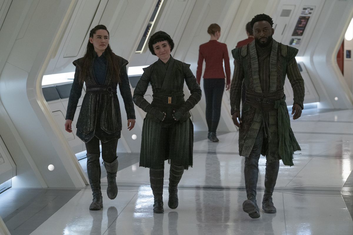 Christina Chong as La’an, Melissa Navia as Ortegas and Babs Olusanmokun as Dr. M’Benga appearing in episode 204 “Among The Lotus Eaters” of Star Trek: Strange New Worlds, streaming on Paramount+, 2023. They are all dressed in Rigellian clothing and walking down an Enterprise hallway. However, only Ortegas wears a hat.