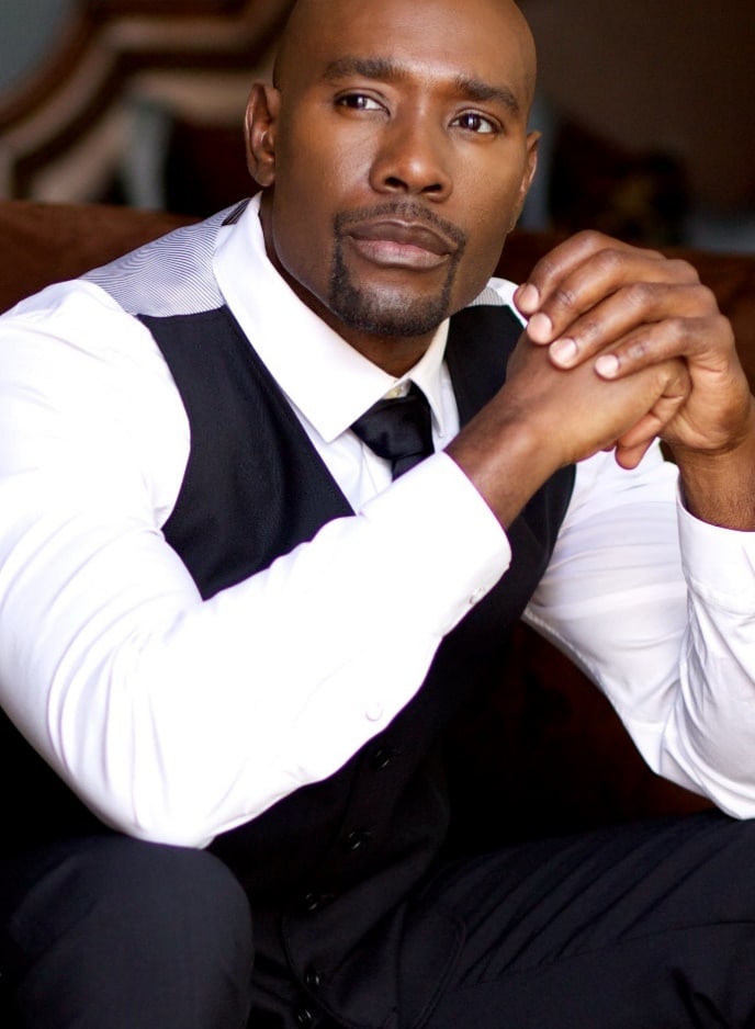 Morris Chestnut, a Black man in a vest and tie, sitting on a couch.
