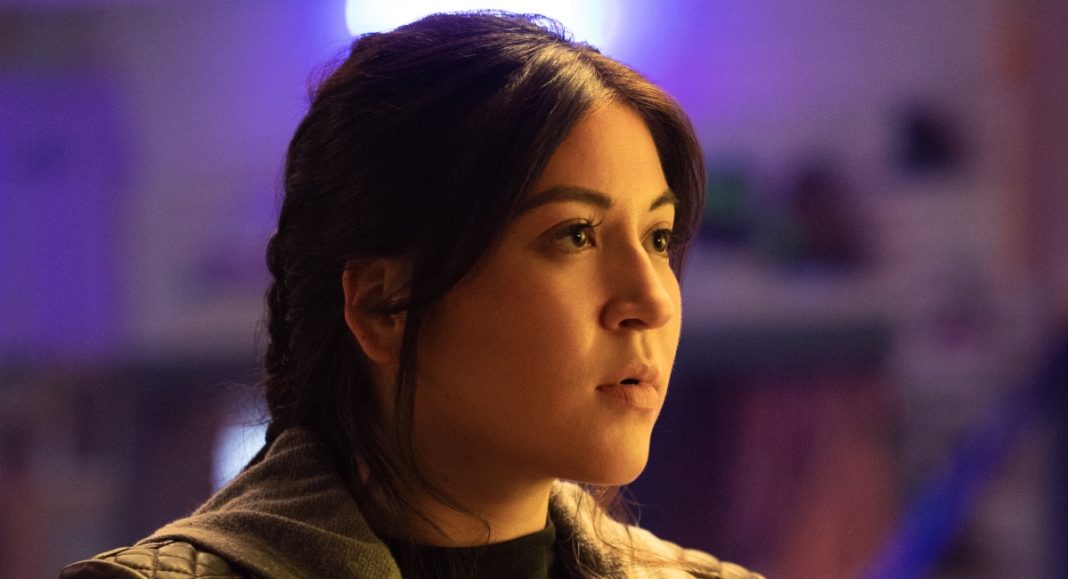 A Native American woman with long, tied-back dark hair looks at something out of frame. She is in front of a purple background.