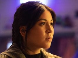 A Native American woman with long, tied-back dark hair looks at something out of frame. She is in front of a purple background.