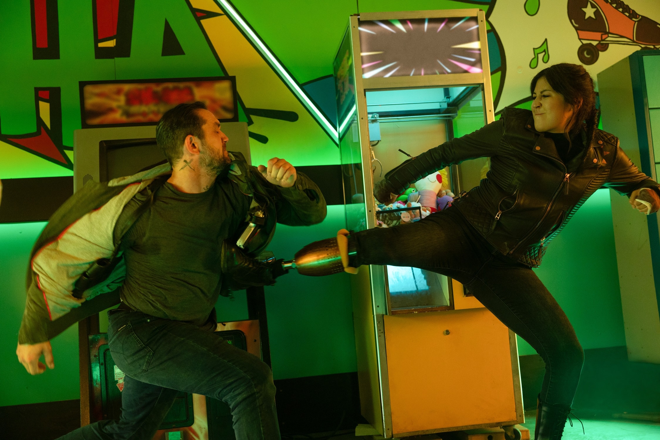 A Native American woman with tied-back long dark hair and a leg prothesis, wearing a black leather jacket and black jeans, kicks a white man with buzzed hair, green jacket, and blue jeans, with her prosthesis. The background is neon, 90's themed, and green-hued.