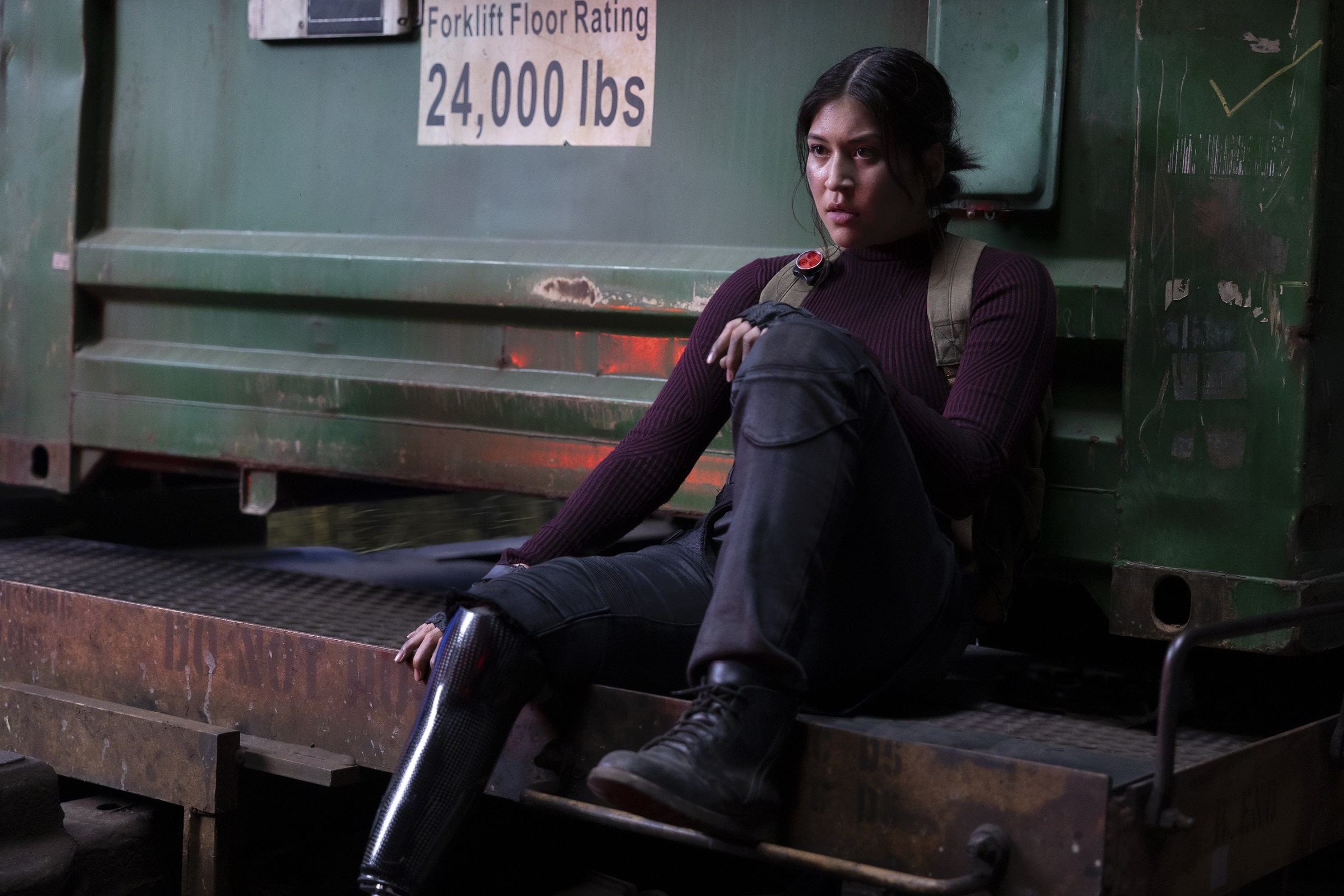 A Native American woman with long, tied-back dark hair and a right-leg prosthesis leans back against a green cargo container on a train. She looks frustrated and/or exhausted.