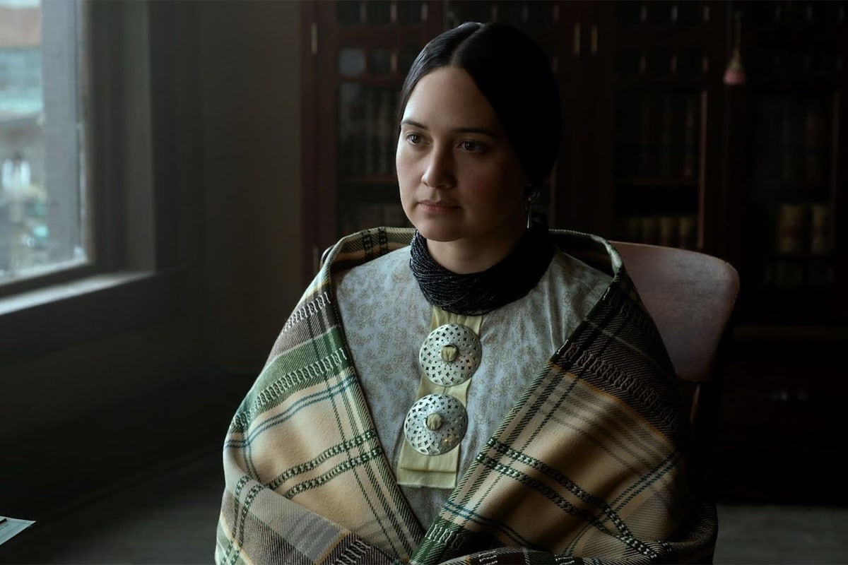 Lily Gladstone, a Native American woman with shoulder-blade-length dark wavy hair, sits in a traditional Osage dress.