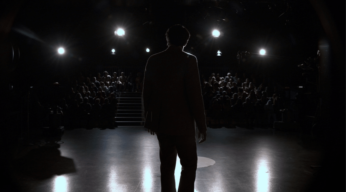 Jack Delroy (played by David Dastmalchian) stands in shadows in front of an audience.