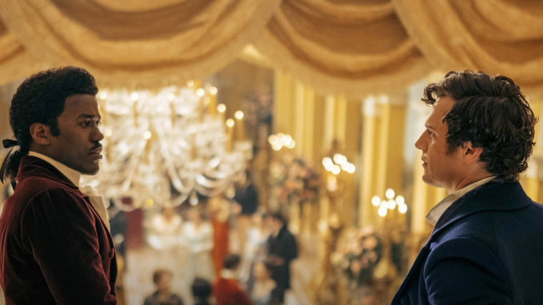 The Doctor (portrayed by Ncuti Gatwa, a black man with his hair tied back, wearing a plum waistcoat and white trousers,) speaks with the mysterious brooding stranger on the balcony (portrayed by Jonathan Groff, a white man with short brown hair wearing a blue waistcoat.)