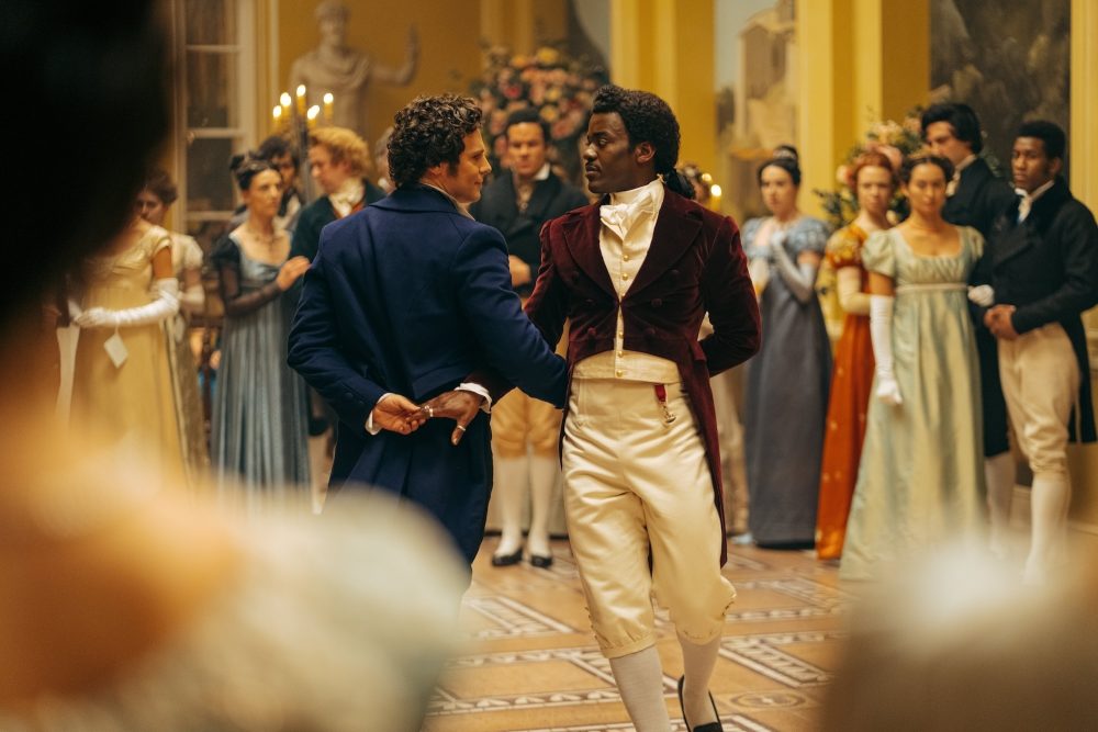 Rogue (a white man with short brown hair and a long blue waistcoat) shares a scandalous dance with the Doctor (a black man with his hair tied back, wearing a plum waistcoat over a white shirt and white trousers.)