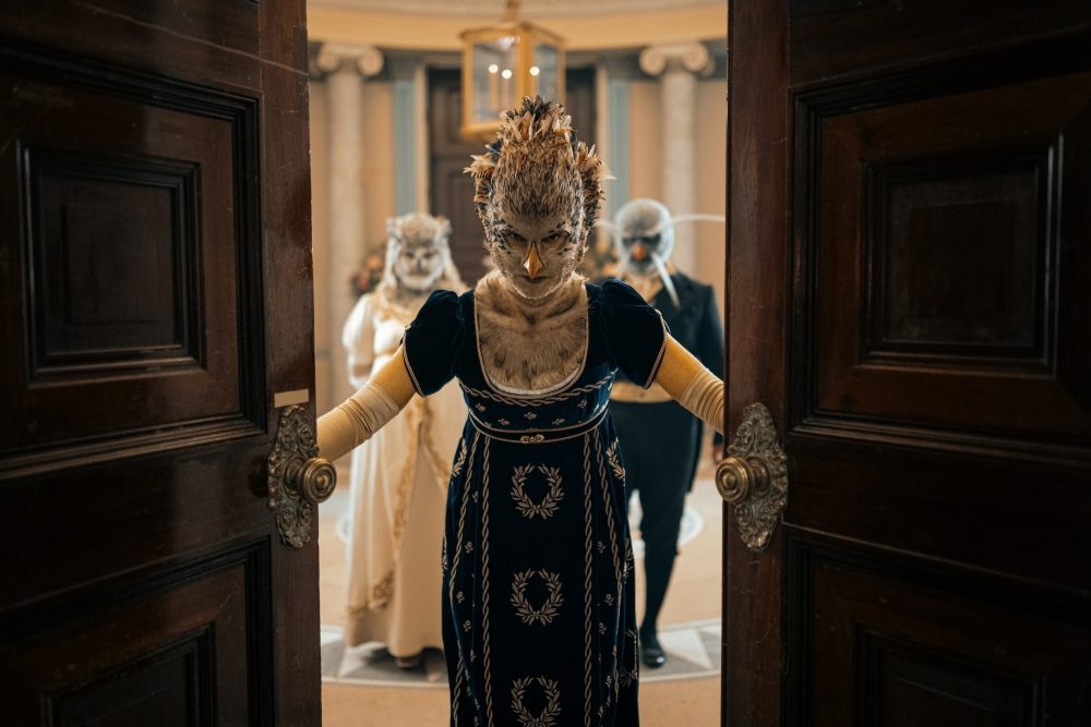 Indira Varma wears a bird-like face and makeup and green dress as she leads the avian alien Chuldur family in their hunt. Two other avian-faced are behind her, one in a white dress, and one in a dark suit.