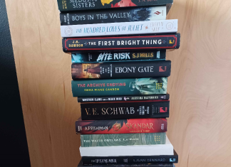 SDCC ’24: The Book Gremlin Guide Several books in a stack against a brown wooden door backdrop