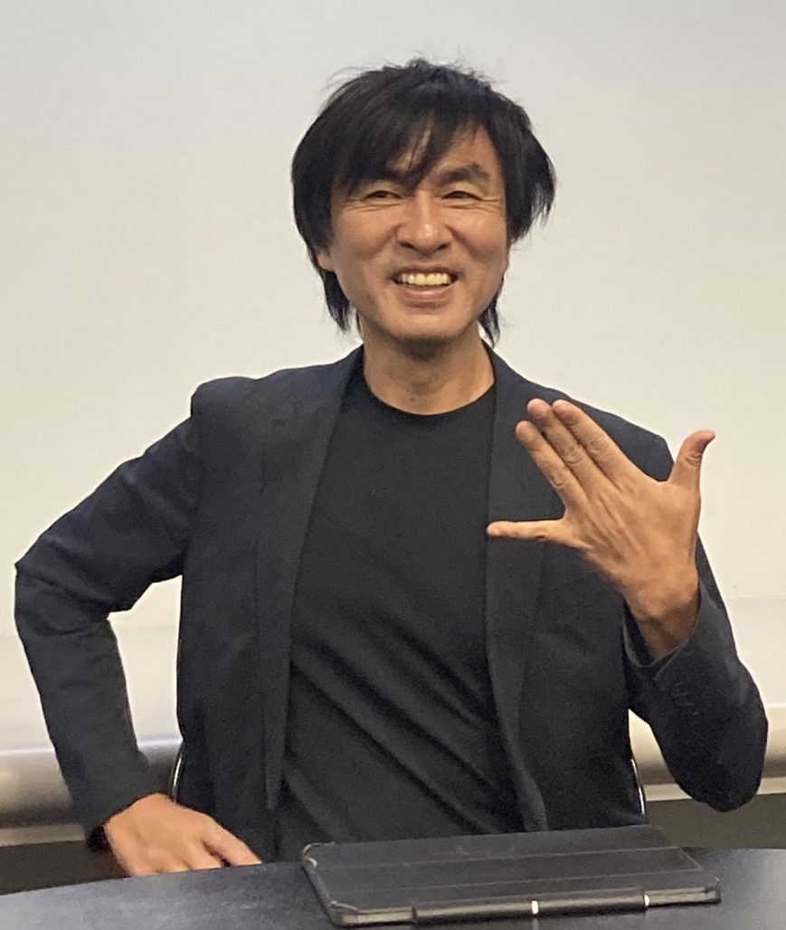 Shoji Kawamori. Smiling man wearing a black jacket and undershirt holds out the pinky and thumb of his left hand, forming a plane symbol.