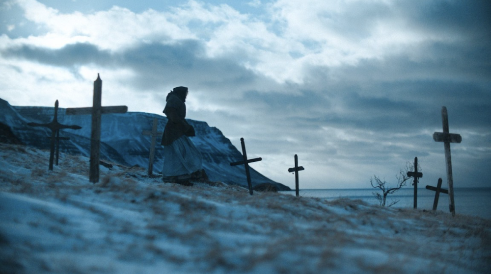 A woman walks along a gray shoreline dotted with graves.