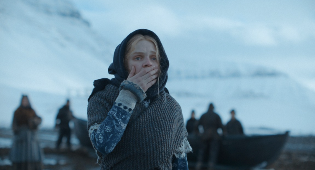 Odessa Young standing in front of men on a bleak white shoreline in The Damned
