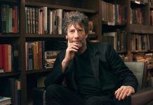 Trafficking lawsuit against Gaiman dismissed Neil Gaiman is sitting on a chair with rows of books behind him