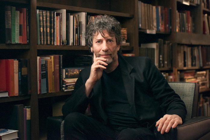 Neil-Gaiman-Masterclass Neil Gaiman is sitting on a chair with rows of books behind him