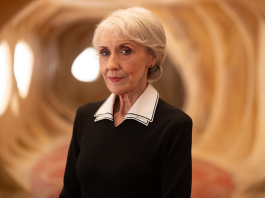 Anita Dobson as Mrs. Flood stands in a well lit beige hallway