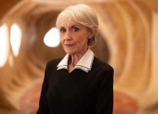 What the Mrs. Flood reveal on DOCTOR WHO means… Anita Dobson as Mrs. Flood stands in a well lit beige hallway