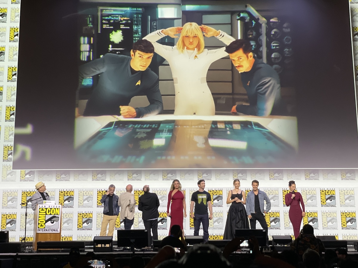 The Strange New Worlds cast and crew on the stage in Hall H at the Star Trek Universe panel, with a picture of Peck, Bush and Dan Jeannotte on set on the screen behind them.