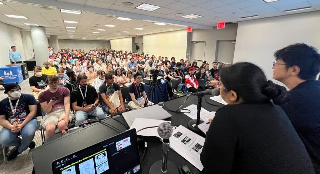 behind shonen jump panel audience photo of panel room showing packed crowd as well as panel moderator and yu saito.