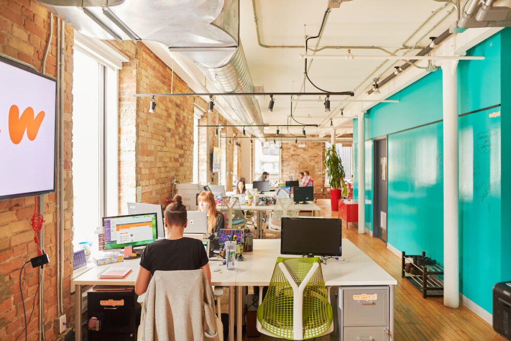 a photograph taken within the Wattpad offices. a woman is at her desk, in front of a computer. her back facing us as a television hangs on the wall to the left with the Wattpad logo on display.
