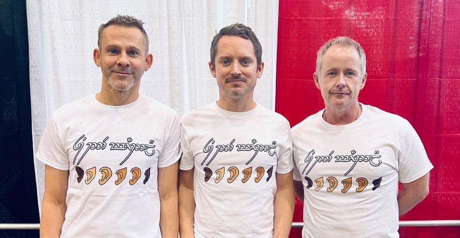 Left to right: Dominic Monaghan, Elijah Wood and Billy Boyd supporting The Rings of Power cast in 2022. The Tengwar script on their shirts, which also features prosthetic ears with different skin colors, says